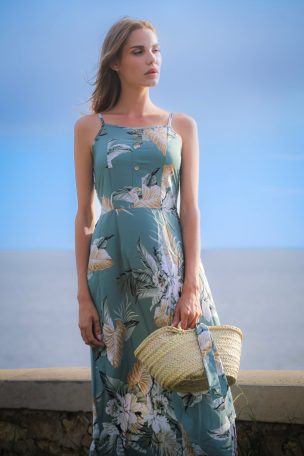 Femme en robe floral, tenant un panier, devant un ciel bleu et une mer calme.