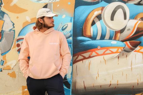 Homme en sweat rose et casquette, posé devant une fresque colorée.