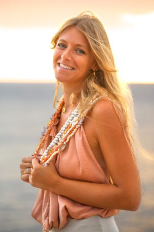 Femme souriante avec un collier, devant un coucher de soleil sur la mer.