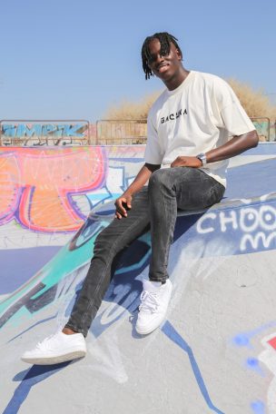 Jeune homme souriant assis sur un mur de skate, portant un t-shirt et des baskets blanches.