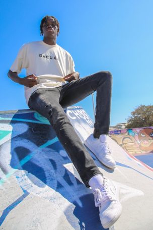 Jeune homme assis sur une rampe de skate, portant un t-shirt et un jean, sous un ciel bleu.