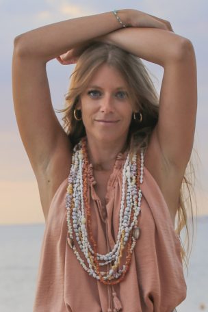 Femme avec des cheveux longs, vêtue d'un top léger et de plusieurs colliers, posant sur la plage.