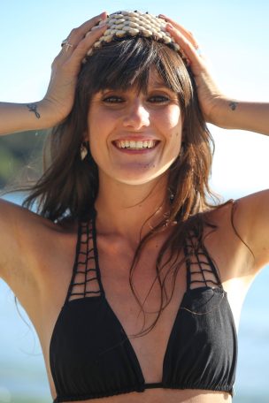 Femme souriante en maillot de bain noir, tenant une couronne avec la mer en arrière-plan.