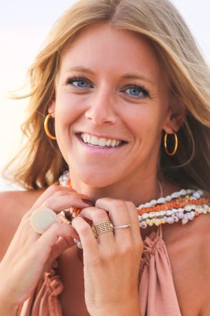 Femme souriante avec des accessoires colorés et des bijoux, posant au bord de la mer.