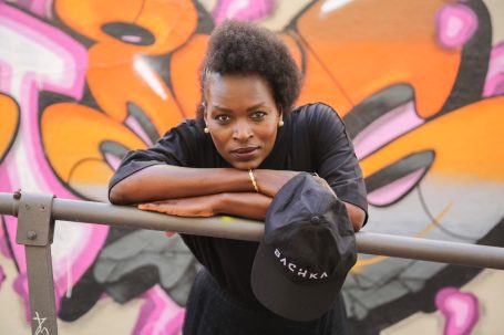 Femme au chapeau noir, posant devant un graffiti coloré.