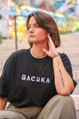 Femme avec un t-shirt noir "BACHKA", assise, regard pensif, arrière-plan coloré.