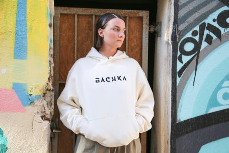 Femme portant un sweat à capuche blanc avec l'inscription «&nbsp;GACHKA&nbsp;», devant un mur coloré.