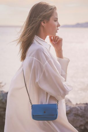 Femme de profil portant une chemise blanche et un sac à main bleu, paysage côtier en arrière-plan.