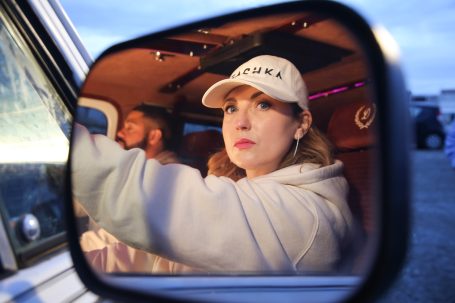 Femme avec une casquette, regardant dans un rétroviseur, avec un homme en arrière-plan.